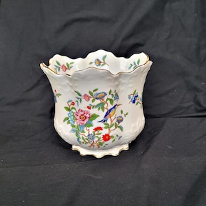 May include: A white ceramic planter with a scalloped rim and a textured surface. The planter is decorated with colourful floral and bird motifs, featuring pink, blue, and yellow accents. The rim has a gold trim.
