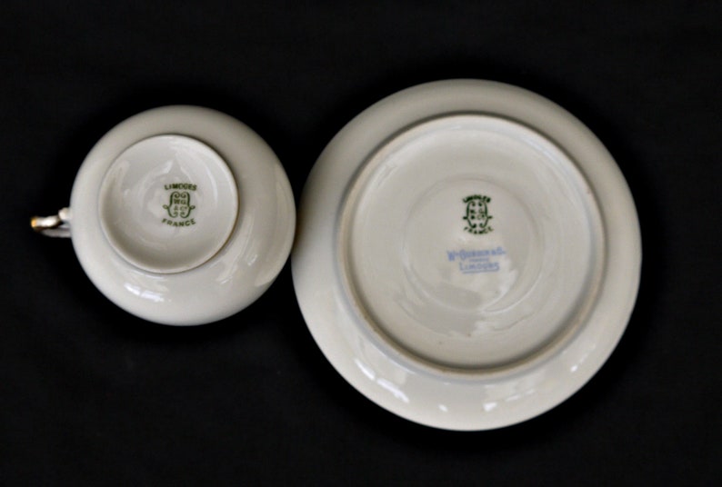 Puede incluir: Un juego de taza y platillo de porcelana blanca con marcas verdes y doradas. El platillo tiene el texto "W. Goebel, Limoges" y la taza tiene el texto "Limoges, France".