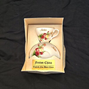 Könnte beinhalten: Ein Fenton China English Fine Bone China Tassen- und Untertassenset in einer Schachtel. Das weiße Porzellan zeigt ein Blumenmuster aus roten und gelben Nelken sowie das Wort Mother in goldener Schrift auf der Tasse und der Untertasse.