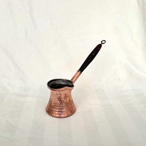 Puede incluir: Una cafetera de cobre con acabado martillado y mango de madera negro. La cafetera tiene un cuerpo redondeado y un pico en el lateral.