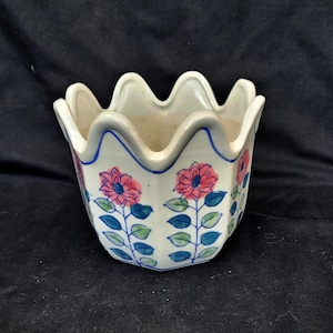 May include: A white ceramic planter with a scalloped rim, decorated with pink and blue floral patterns. The planter features a wavy edge and blue trim, suitable for small plants or flowers.
