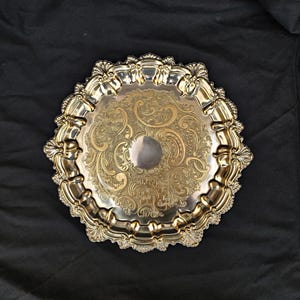 May include: A round, silver-toned serving tray with an ornate, embossed floral design. The tray has a scalloped edge with decorative detailing. The center of the tray has a circular indentation. The tray is displayed against a black background.