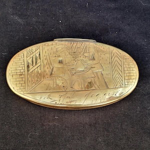 May include: Oval brass antique box with an engraved scene of people at a table, possibly a tavern. The box has a hinged lid and is decorated with brickwork and a window. The bottom of the box has engraved text.