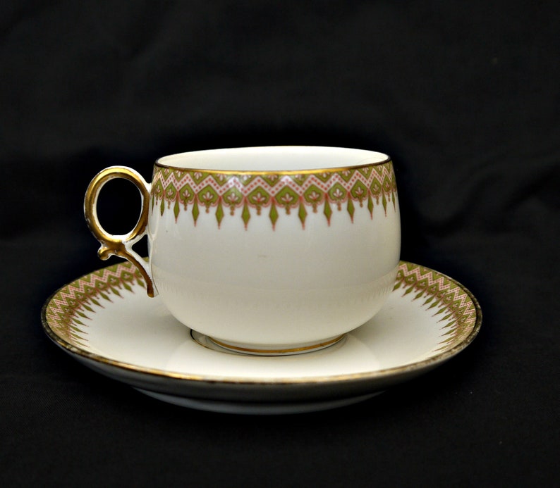 Puede incluir: Una taza de t&eacute; y un platillo blancos con un borde dorado y un patr&oacute;n floral verde y rosa.