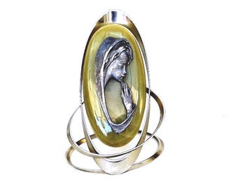 May include: A silver-toned ring featuring an oval-shaped, yellow-tinted glass centerpiece. Inside the glass is a detailed silver relief of a woman in prayer. The ring has a unique, looping design around the central oval.