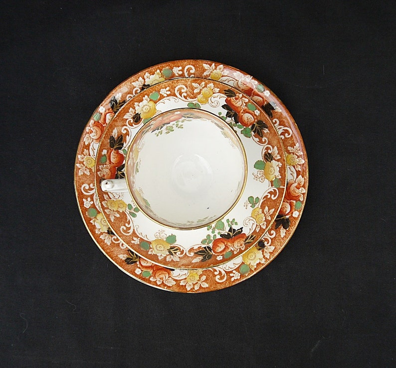 May include: A white teacup with a floral pattern sits on a saucer with a similar floral pattern. The saucer has a brown background with orange, red, and yellow flowers.