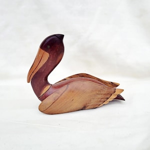May include: A wooden pelican sculpture with brown and light brown wood grain. The pelican's head is turned to the side and its wings are spread out.