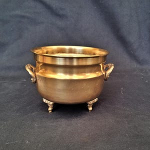 Peut inclure: Un petit chaudron en laiton antique avec trois pieds ornés et deux poignées décoratives. Le chaudron a un corps arrondi et un large bord plat. Le laiton a une teinte dorée chaude, suggérant l'âge et l'utilisation. Le chaudron est probablement utilisé à des fins décoratives.