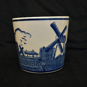 May include: A white ceramic planter with a blue and white design of a windmill and houses. The planter has a blue rim and a blue and white pattern around the bottom.