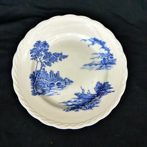 May include: A white ceramic plate with a blue and white landscape design. The design features a river, a bridge, a castle, trees, and a boat with two people in it.