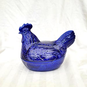 Pode incluir: Um recipiente de cerâmica azul cobalto em forma de galinha. A tampa e a base apresentam detalhes texturizados. A crista e as penas da cauda da galinha são claramente definidas. Esta peça decorativa é provavelmente usada para armazenamento ou exibição.