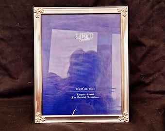 May include: Silver-colored picture frame with floral corner accents. The frame is 8 x 10 inches and has the text "SHUDEHILL" and "Lacquer Coated For Tarnish Resistance". The frame is on a dark brown background.