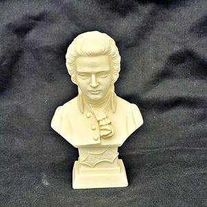 Può includere: Un busto in marmo bianco di Wolfgang Amadeus Mozart su una base bianca. Il busto è dettagliato e mostra il viso e i capelli di Mozart. La base ha l'iscrizione "W.A. Mozart".