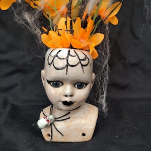 May include: A ceramic doll head planter featuring a painted black spiderweb on the forehead, dark eyeliner, and black lips. A small skull charm with red eyes hangs from the neck. The head is filled with bright orange artificial flowers and white cobwebs.