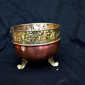 May include: A round copper bowl featuring a wide brass rim embossed with a continuous relief of figures in motion. The bowl is supported by three ornate brass feet with a curved design, contrasting with the smooth, polished copper body.