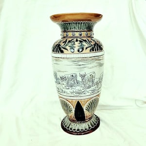 May include: A decorative ceramic vase with a wide base and flared top. The vase features a landscape scene with cattle, and is adorned with floral and geometric patterns in brown, green, and cream. The vase is approximately 40 cm tall.