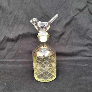 Puede incluir: Una botella de vidrio transparente con un tapón en forma de pájaro. La botella tiene un tinte amarillo y un patrón de diamantes grabado en el vidrio. El tapón de pájaro también es de vidrio transparente.