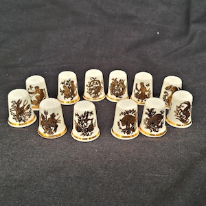 Könnte beinhalten: Eine Sammlung von zwölf weißen Porzellan-Fingerhüten mit goldenen Rändern und detaillierten braunen Illustrationen. Jeder Fingerhut zeigt ein anderes Design, möglicherweise Tierkreiszeichen oder mythologische Figuren.
