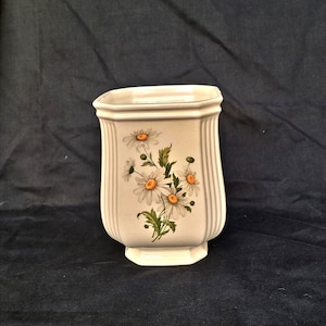 Puede incluir: Un jarrón de cerámica blanco con un diseño floral de margaritas. El jarrón tiene forma rectangular con detalles acanalados verticales y una base acampanada. El diseño de la margarita presenta pétalos blancos y centros amarillos.