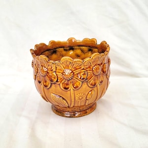 May include: A brown ceramic flower pot with a repeating floral pattern. The pot has a scalloped rim and a small pedestal base. The interior is dark brown. The pot is decorated with embossed flowers and leaves.