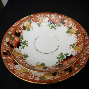 May include: A set of two vintage ceramic plates with a floral design. The plates feature a brown background with gold accents and a white center. The floral design includes roses, leaves, and swirls.