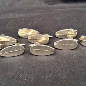 May include: A collection of ten small, metallic, teardrop-shaped objects with a smooth, reflective finish. Each piece features a distinct horizontal seam running across the centre of its rounded, elongated form, creating a uniform set of components.