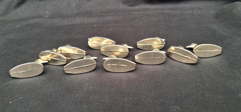 May include: A collection of ten small, metallic, teardrop-shaped objects with a smooth, reflective finish. Each piece features a distinct horizontal seam running across the centre of its rounded, elongated form, creating a uniform set of components.