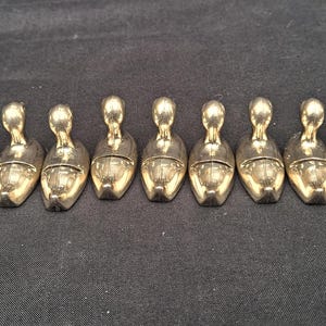 May include: A set of twelve identical metallic shoe-shaped objects arranged in a horizontal row. Each piece features a smooth, reflective gold-toned finish, a rounded toe, and a small upright handle or loop extending from the heel section.