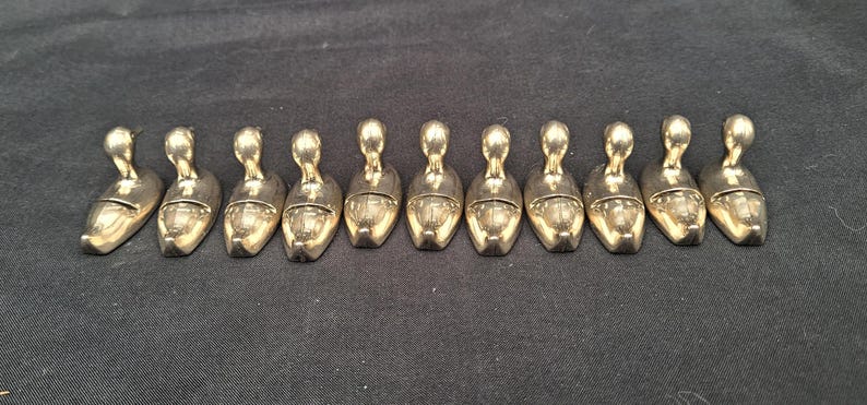 May include: A set of twelve identical metallic shoe-shaped objects arranged in a horizontal row. Each piece features a smooth, reflective gold-toned finish, a rounded toe, and a small upright handle or loop extending from the heel section.