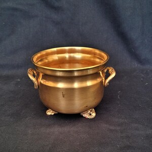 May include: A small, round, brass-coloured cauldron with two ornate handles and three small feet. The cauldron has a wide rim and a smooth, polished surface. The design is simple yet elegant, with a vintage or antique aesthetic.