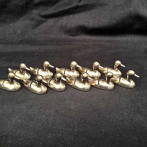May include: A collection of ten small, polished brass duck figurines arranged in two rows. Each duck features a smooth, metallic gold finish with a simple, stylised form, including a defined head, neck, and body, all facing toward the right side.