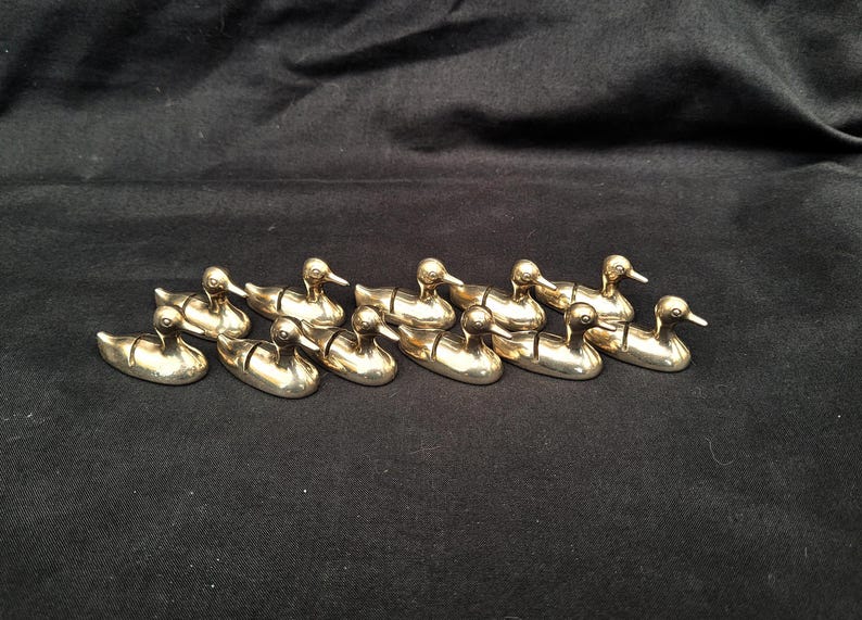 May include: A collection of ten small, polished brass duck figurines arranged in two rows. Each duck features a smooth, metallic gold finish with a simple, stylised form, including a defined head, neck, and body, all facing toward the right side.
