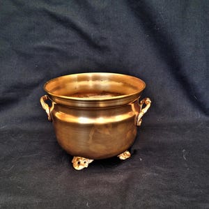Può includere: Una piccola ciotola in ottone in stile antico con un corpo arrotondato, due manici ornati e tre piedini decorativi. La ciotola ha una finitura dorata lucida e poggia su uno sfondo scuro. Questa ciotola decorativa è adatta a vari usi.