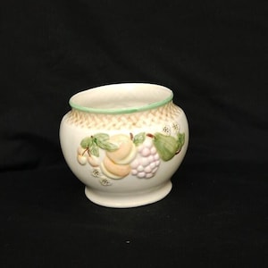 May include: A white ceramic bowl featuring a raised, textured basket-weave pattern around the rim with a light green edge. The exterior is decorated with embossed, hand-painted fruit motifs including peaches, grapes, a pear, and small white flowers.