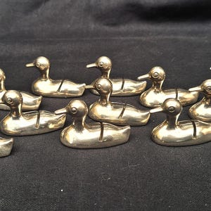 May include: A collection of ten small, metallic, silver-toned duck-shaped figurines. Each duck features a smooth, polished surface and a distinct vertical slit cut into the back, designed to hold small items like place cards or notes.