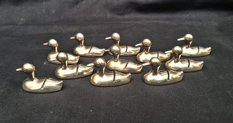 May include: A collection of ten small, metallic, silver-toned duck-shaped figurines. Each duck features a smooth, polished surface and a distinct vertical slit cut into the back, designed to hold small items like place cards or notes.