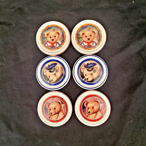 May include: Six small, round decorative plates with a teddy bear design. The plates feature a white background with a colored border and a central image of a teddy bear in various outfits. The borders are in orange, blue, and red.