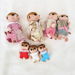 Puede incluir: Colecci&oacute;n de siete figuras de suricatos vestidos con varios atuendos. Los suricatos llevan vestidos, monos y chaquetas en tonos rosa, beige, azul y verde. Las figuras tienen pelaje marr&oacute;n y est&aacute;n posadas sobre una superficie blanca.