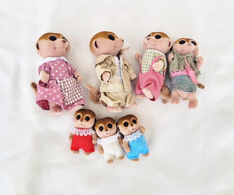 Puede incluir: Colecci&oacute;n de siete figuras de suricatos vestidos con varios atuendos. Los suricatos llevan vestidos, monos y chaquetas en tonos rosa, beige, azul y verde. Las figuras tienen pelaje marr&oacute;n y est&aacute;n posadas sobre una superficie blanca.