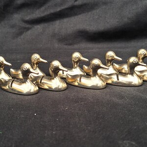 May include: A collection of nine small, polished gold-toned metal duck figurines arranged in a row. Each duck features a smooth, reflective surface, a defined beak, and a small slit on its back, designed to hold items in an upright position.