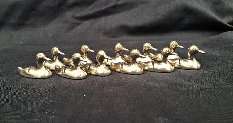 May include: A collection of nine small, polished gold-toned metal duck figurines arranged in a row. Each duck features a smooth, reflective surface, a defined beak, and a small slit on its back, designed to hold items in an upright position.
