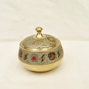 Puede incluir: Un pequeño recipiente redondo con tapa y diseño ornamentado. El recipiente es principalmente dorado, con una banda plateada con detalles florales rojos. La tapa tiene un pomo decorativo y patrones florales en relieve.