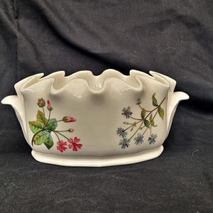 Puede incluir: Un cuenco de cerámica blanco con borde ondulado y asas. El cuenco está decorado con diseños florales en rosa, azul y verde. El cuenco es probablemente para fines decorativos o para servir.