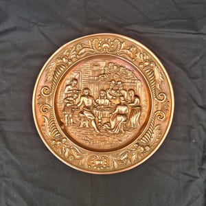 Könnte beinhalten: Eine kupferfarbene Zierplatte mit einer detaillierten Reliefszene von Menschen, die sich um einen Tisch versammeln. Die Platte hat einen verzierten floralen Rand und einen leicht erhöhten Rand. Die Szene zeigt ein Treffen von Menschen.
