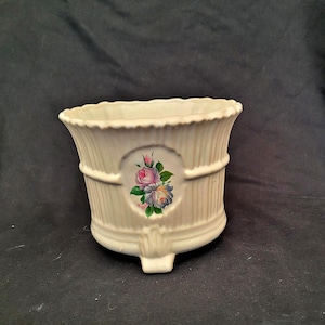 May include: Cream-coloured ceramic planter with a ribbed design and a floral medallion. The planter features a scalloped rim and a decorative painted design of pink, purple, and blue roses. The planter has a small foot.