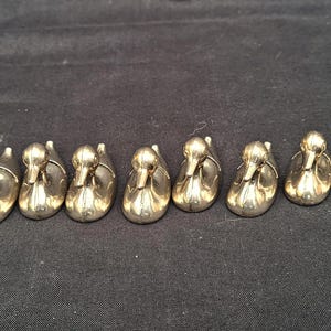 May include: A collection of eleven small, polished gold-tone metal duck figurines arranged in a horizontal row. Each duck is depicted in a seated or swimming position with a smooth, reflective surface and a simple, stylised form facing forward.