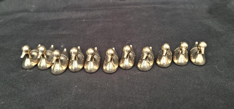 May include: A collection of eleven small, polished gold-tone metal duck figurines arranged in a horizontal row. Each duck is depicted in a seated or swimming position with a smooth, reflective surface and a simple, stylised form facing forward.
