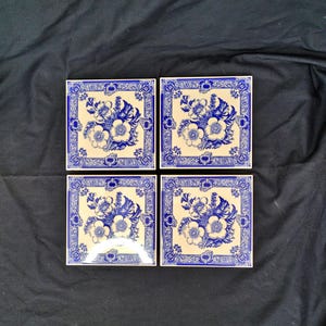 May include: Four square ceramic tiles with a floral design. Each tile has a cream background with a blue floral arrangement and a blue border. The flowers are white with blue accents.