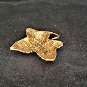 Könnte beinhalten: Ein goldfarbenes, blattförmiges Dekorationsobjekt. Das Objekt hat detaillierte Adern und eine Schlaufe oben. Das Blatt liegt auf einer dunklen Oberfläche. Dieser Artikel könnte als Dekorationselement oder Schmuckkomponente verwendet werden.