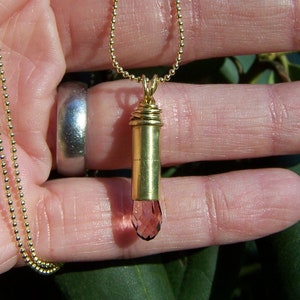 May include: A gold chain necklace with a bullet casing pendant filled with a pink gemstone.